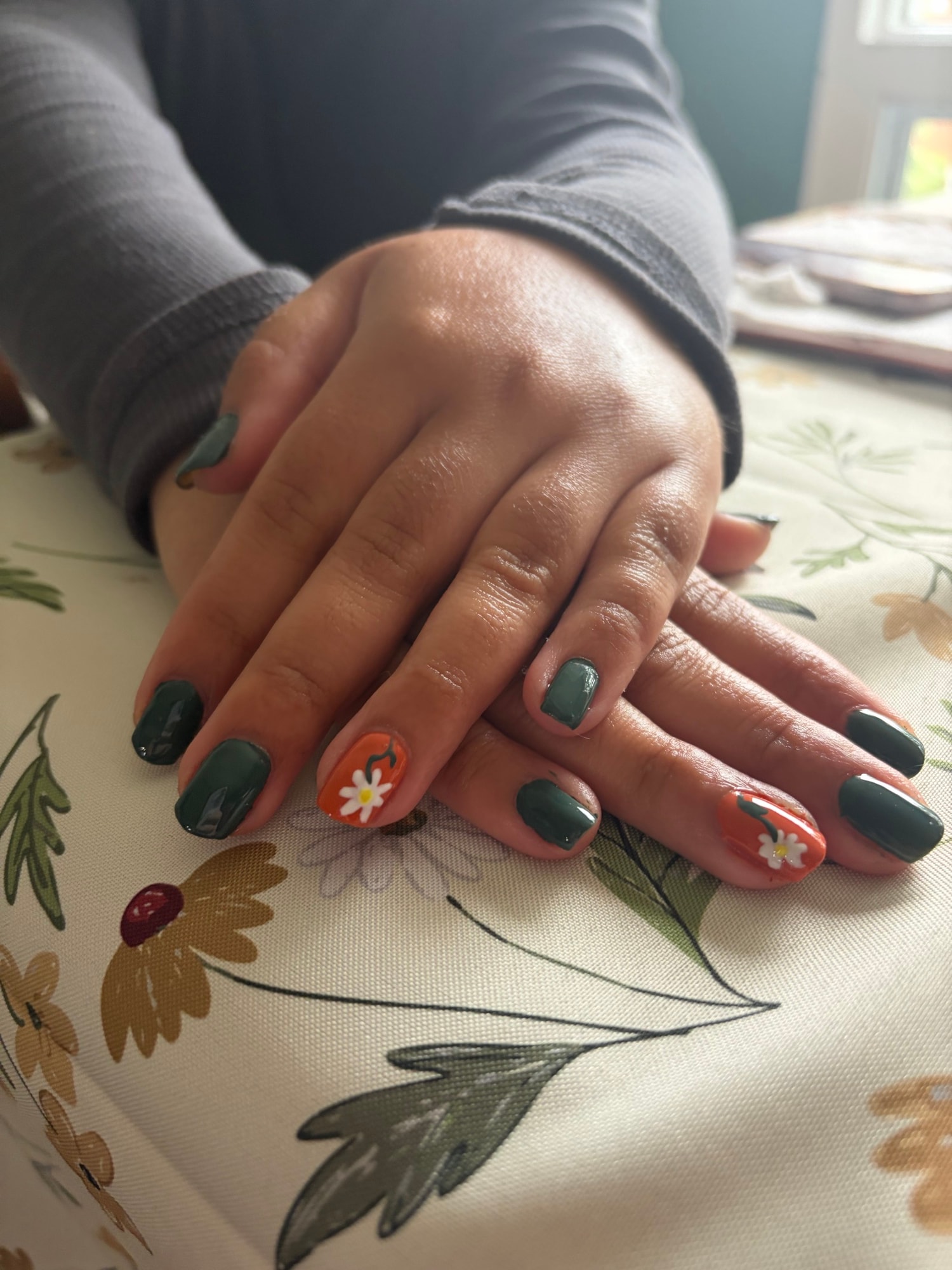 Mains aux ongles courts vert forêt, avec deux ongles orange ornés d'une marguerite peinte